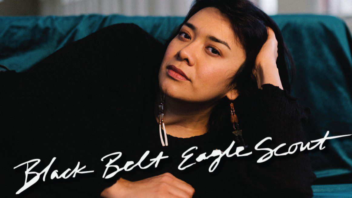 An Indigenous woman lays down and poses with a hand in her hair. Text reads "Black Belt Eagle Scout."
