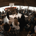 A person breakdances under a large pavilion in the middle of a large crowd of other breakdancers and a DJ.