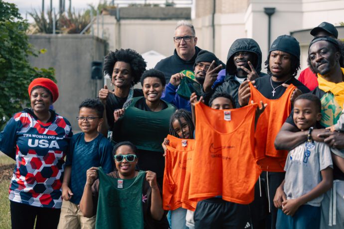 A mostly Black big group of people of all ages smile and hold up soccer jerseys.