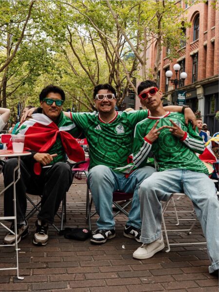 Three soccer fans wearing Mexico soccer jerseys and shades with their arms around each other in Pioneer Square.