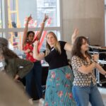 Three women joyfully dance in a sunny room