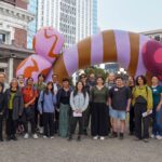 A group of 18 artists stand on the King Street Plaza in front of a large colorful sculpture