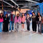 Seven teens and one adult woman pose together under a large suspended artwork of a Black woman calmly flying.