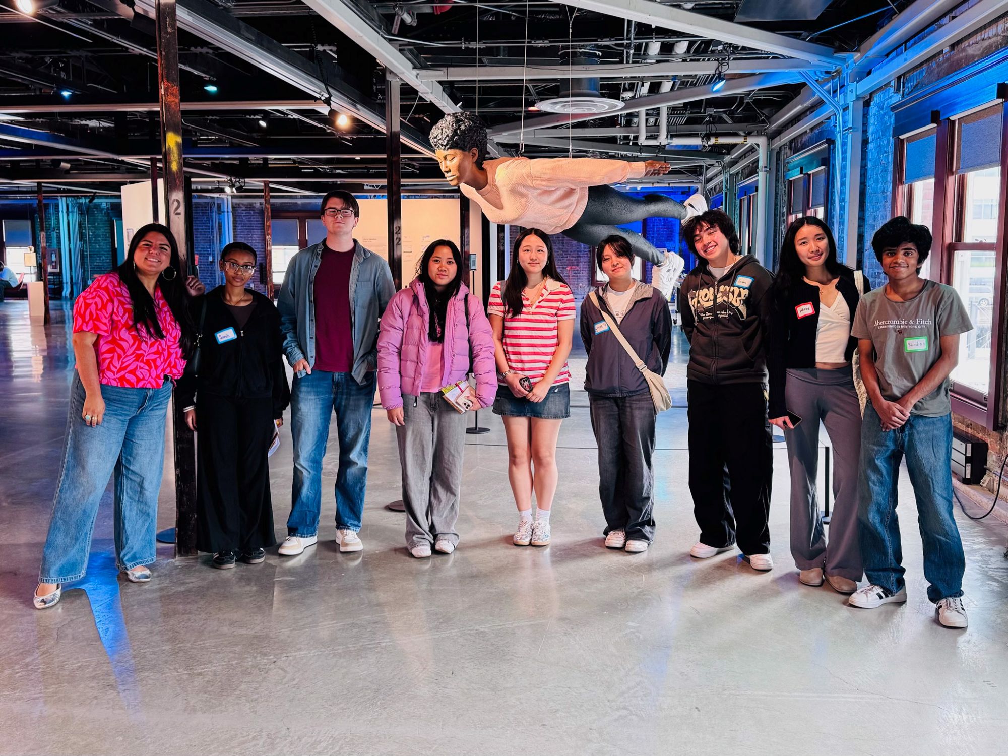 Seven teens and one adult woman pose together under a large suspended artwork of a Black woman calmly flying.