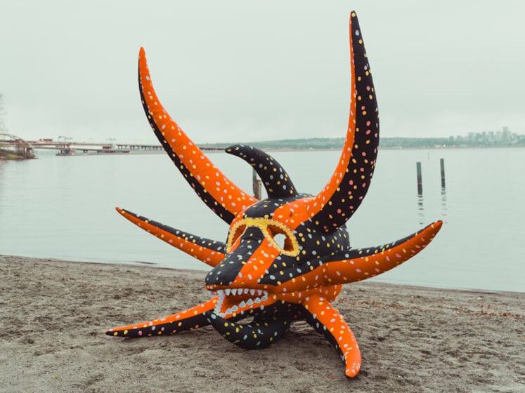 A orange and black with white polka dot imagined sea creature wearing sunglasses on the beach with water in the background.