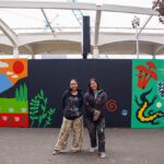 Christina and Stevie pose in paint spattered clothing in front of their mural. On the left is a panel depicting mountains in a natural scene. On the rights is a close-up of things on the ground: a lizard, pine cone, mushrooms, and leaves.